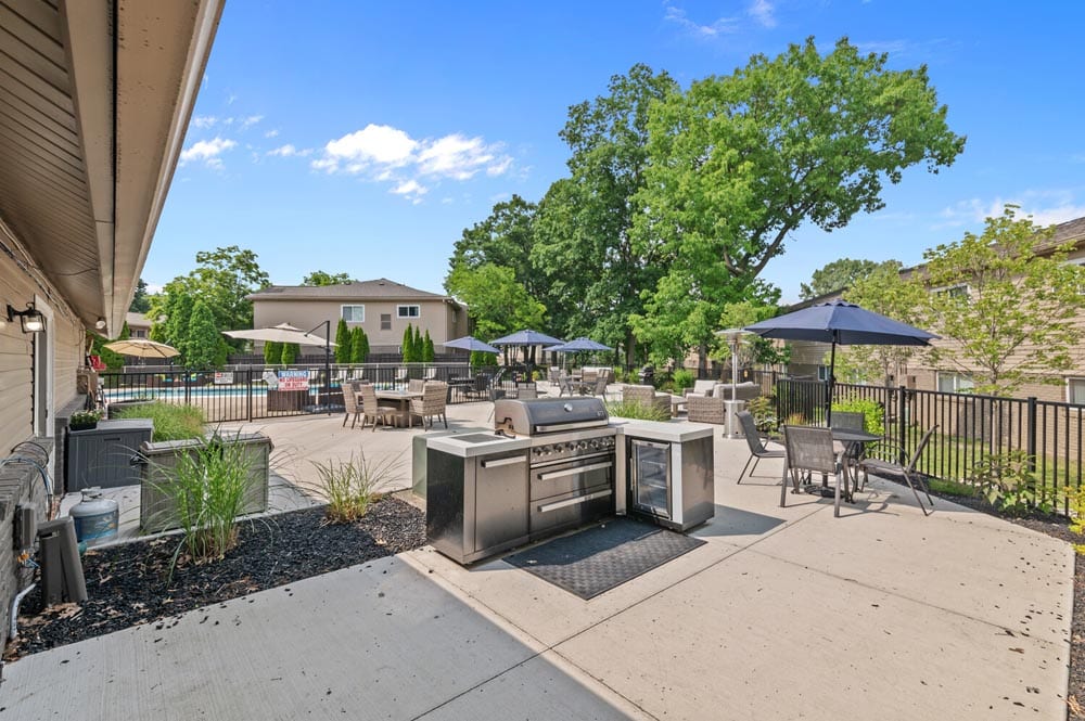 Outdoor patio with grill, tables, and umbrellas beside a pool on a sunny day.