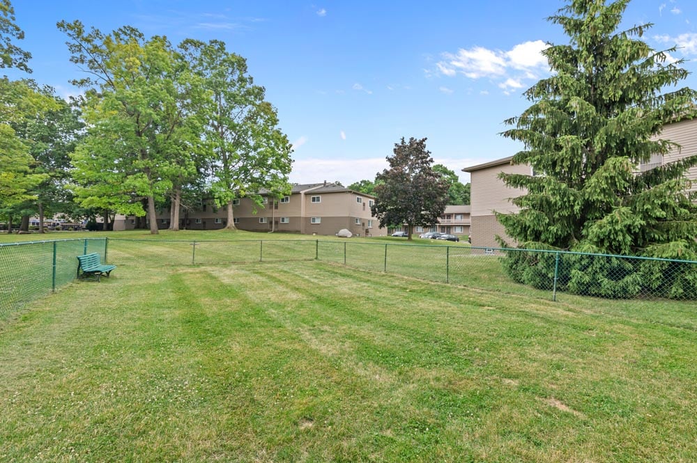 Open grassy area with trees and a fence near residential buildings under a blue sky.
